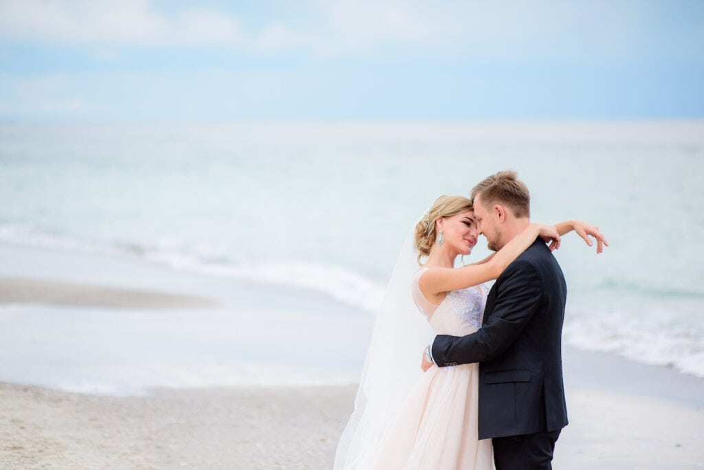 Dreamy wedding couple hug each other tender standing on the beach with sea waves splashing behind them